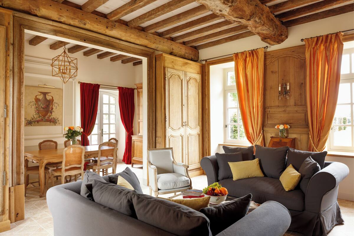 Un ampio soggiorno con soffitto in legno, un divano grigio, un'area pranzo con tavolo e sedie in legno e ampie finestre con tende.