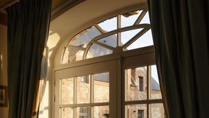 A room with a large arched window, a wooden ceiling, and a view of a stone building outside. - Le Domaine de Ravenoville (Sainte-Mere-Eglise)