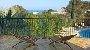 A balcony with two wooden lounge chairs, a small table, and a view of a swimming pool and ocean. - La Maison du Midi by Interhome (Les Issambres)