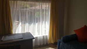 A room with a desk and chair, a blue sofa with a red pillow, and a window with yellow curtains. - Pachigomo Guest House (Goromonzi)