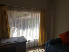 Una habitación con un escritorio y una silla, un sofá azul con un cojín rojo, y una ventana con cortinas amarillas.