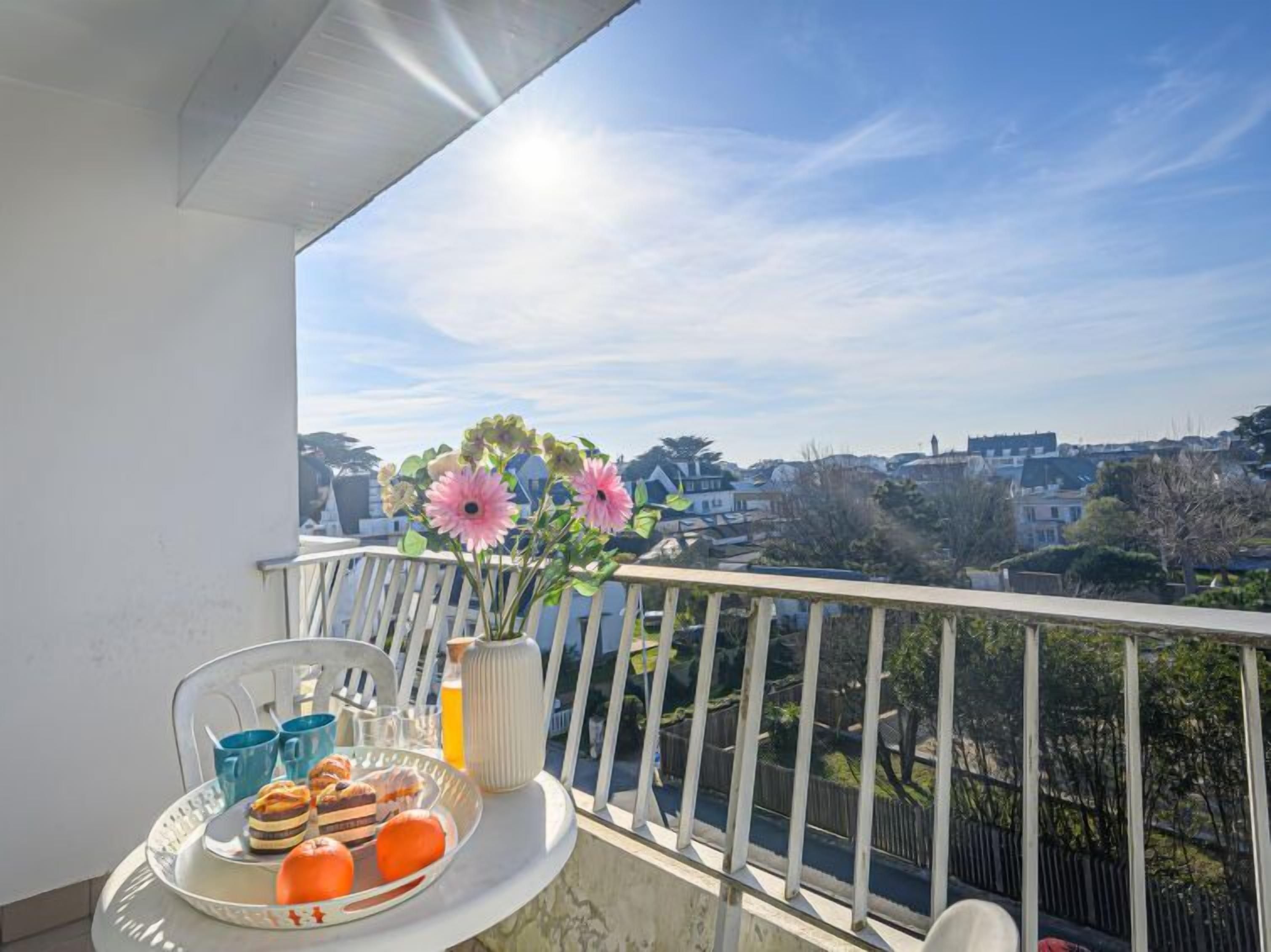 A balcony with a table set for breakfast, including pastries, fruit, and a drink, overlooking a residential area with houses and trees.