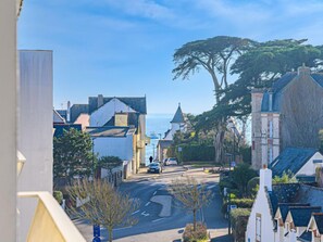 Une rue côtière bordée de bâtiments à l’architecture traditionnelle, offrant une vue dégagée sur la mer et abritant un arbre remarquable.