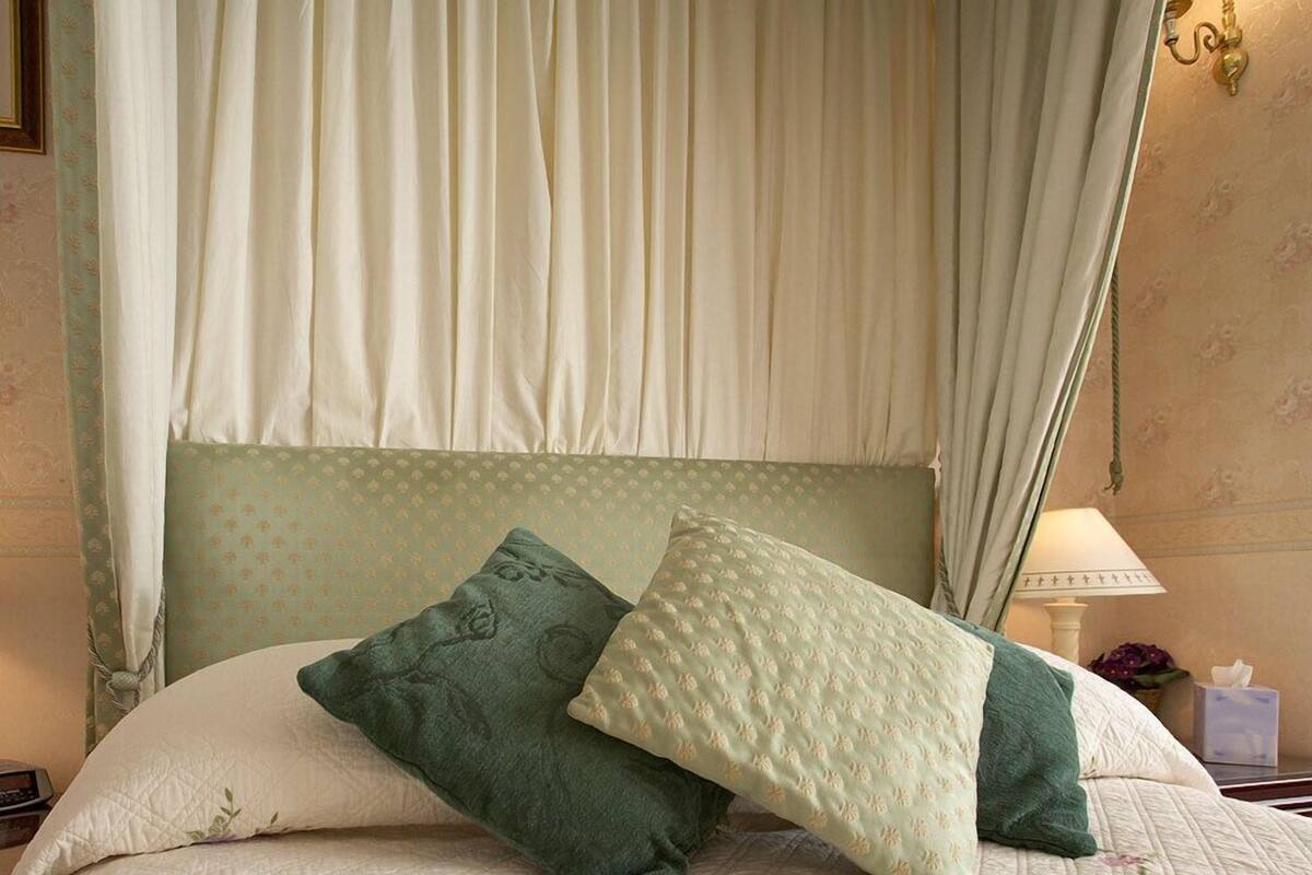 Una cama con dosel, dos almohadas, una mesita de noche con lámpara y una pared con estampado floral.