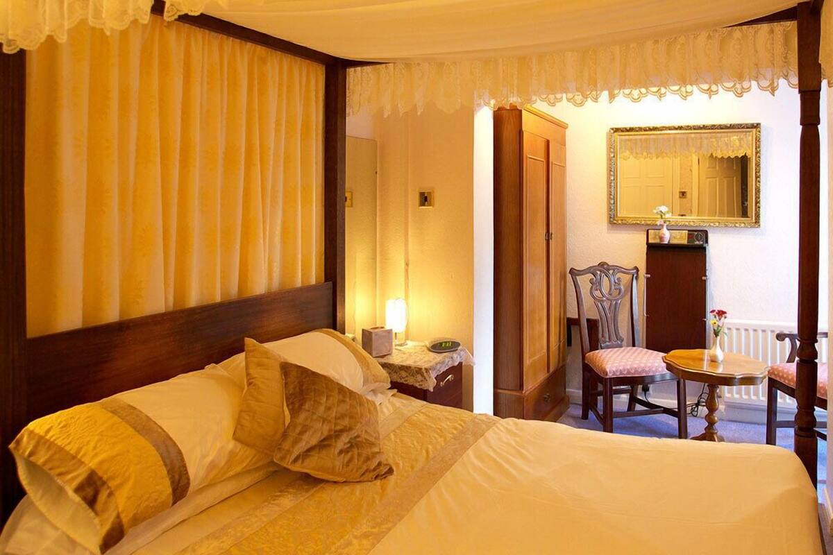 Habitación de hotel con una cama grande, cabecera de madera, mesita de noche con lámpara, una silla, una mesa pequeña con un jarrón y un espejo en la pared.