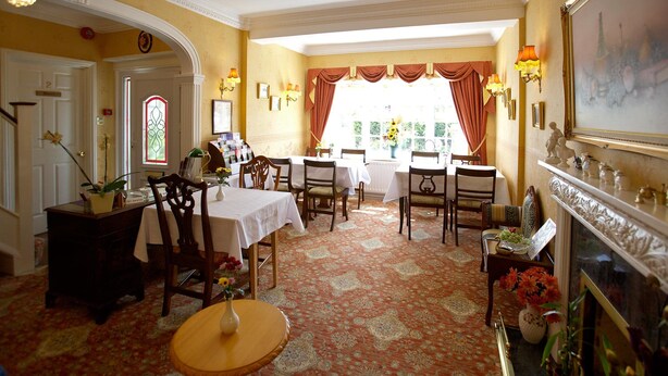 A dining area with tables and chairs, a fireplace, and a large window with curtains. - Moonraker House (Stratford-upon-Avon)