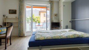 Une chambre avec un lit, un bureau, une chaise, une télévision et un balcon offrant une vue sur les montagnes et un immeuble.