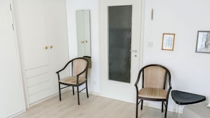 Une pièce comprenant deux chaises en bois, un canapé gris et une petite table noire. On y trouve également une armoire blanche avec un miroir et une porte dotée d’un panneau noir.