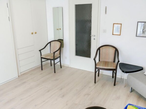 A room with two wooden chairs, a grey sofa, and a small black table. There is a white wardrobe with a mirror and a door with a black panel.