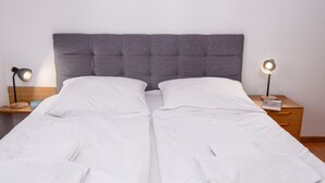 Ein Doppelbett mit weißen Bettwäsche, ein grauer möbelbezogener Kopfteil und zwei Nachttische mit Lampen.