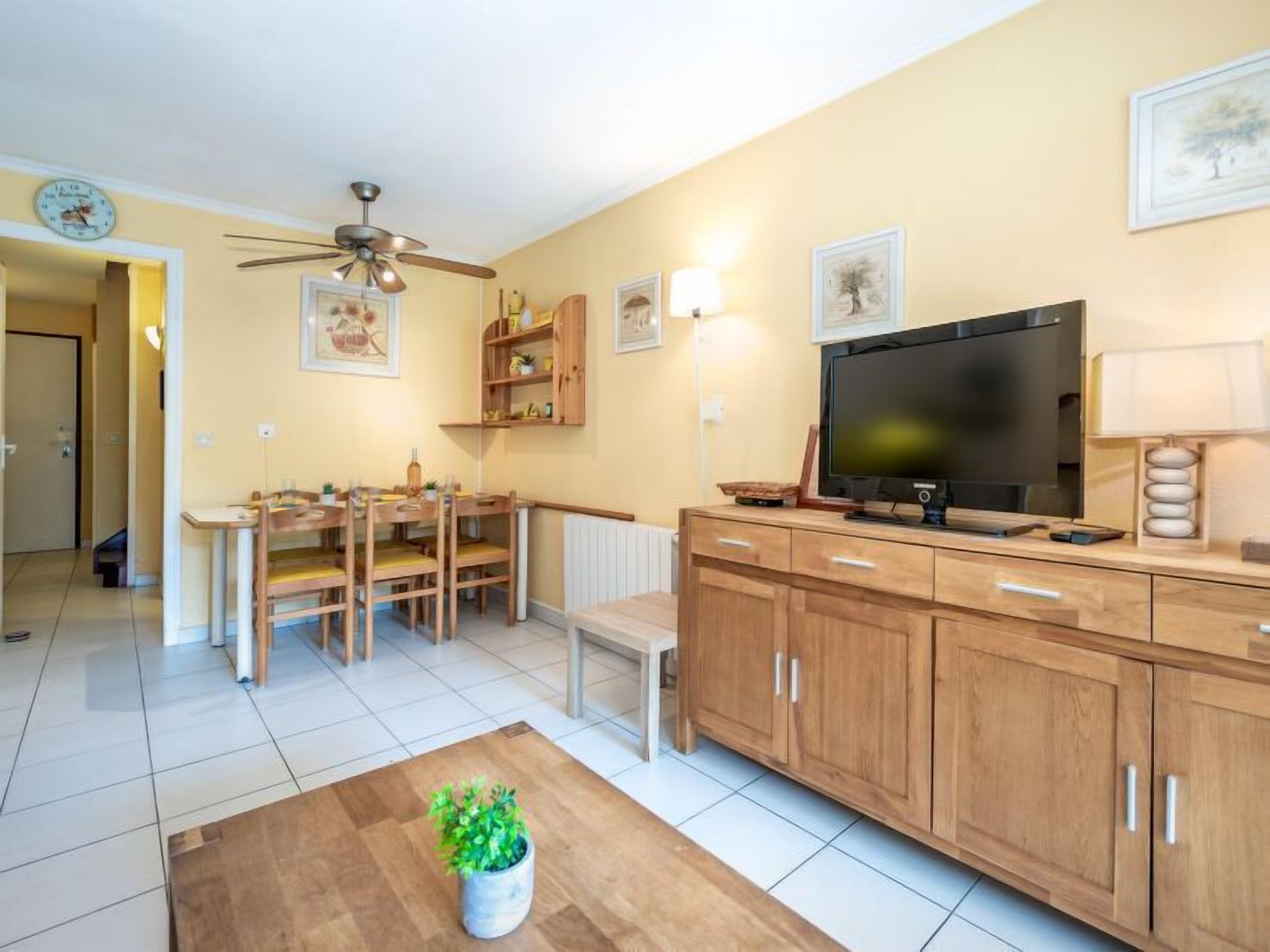 Un salon moderne comprenant un coin repas, des meubles en bois, un ventilateur de plafond, un téléviseur et des œuvres d’art accrochées aux murs.