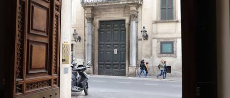 Vista de una calle con una puerta de madera grande, una motocicleta estacionada al lado, y peatones caminando por la acera.