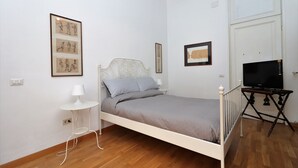 A bedroom with a bed, a small table with a lamp, a TV on a stand, and framed artwork on the wall. - Trevi suites B&B (Rome)