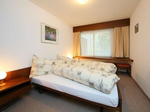 Ein Einzelbett mit weißer Matratze und passenden Kissen, ein Nachttisch mit Lampe, ein Fenster mit Vorhängen und ein gerahmtes Bild an der Wand.