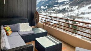 Un balcon agrémenté de meubles en osier, d’une table en verre et offrant une vue sur des montagnes enneigées.