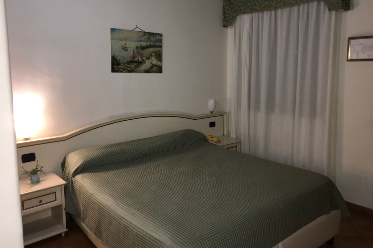 Une chambre avec un lit simple, une table de chevet avec une lampe, un tableau encadré au mur et une fenêtre avec des rideaux.