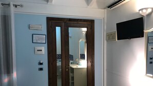 A hotel room with a wooden door, a desk with a chair, a flat-screen TV mounted on the wall, and a bathroom visible through the door. - Motel 24h (Lusciano)