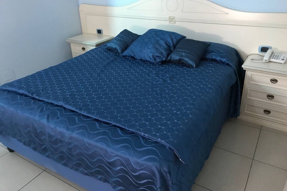Un lit bien fait, avec une couvre-lit bleue à motifs et des oreillers, une tête de lit blanche et une table de chevet sur laquelle se trouvent un téléphone et une petite lampe.