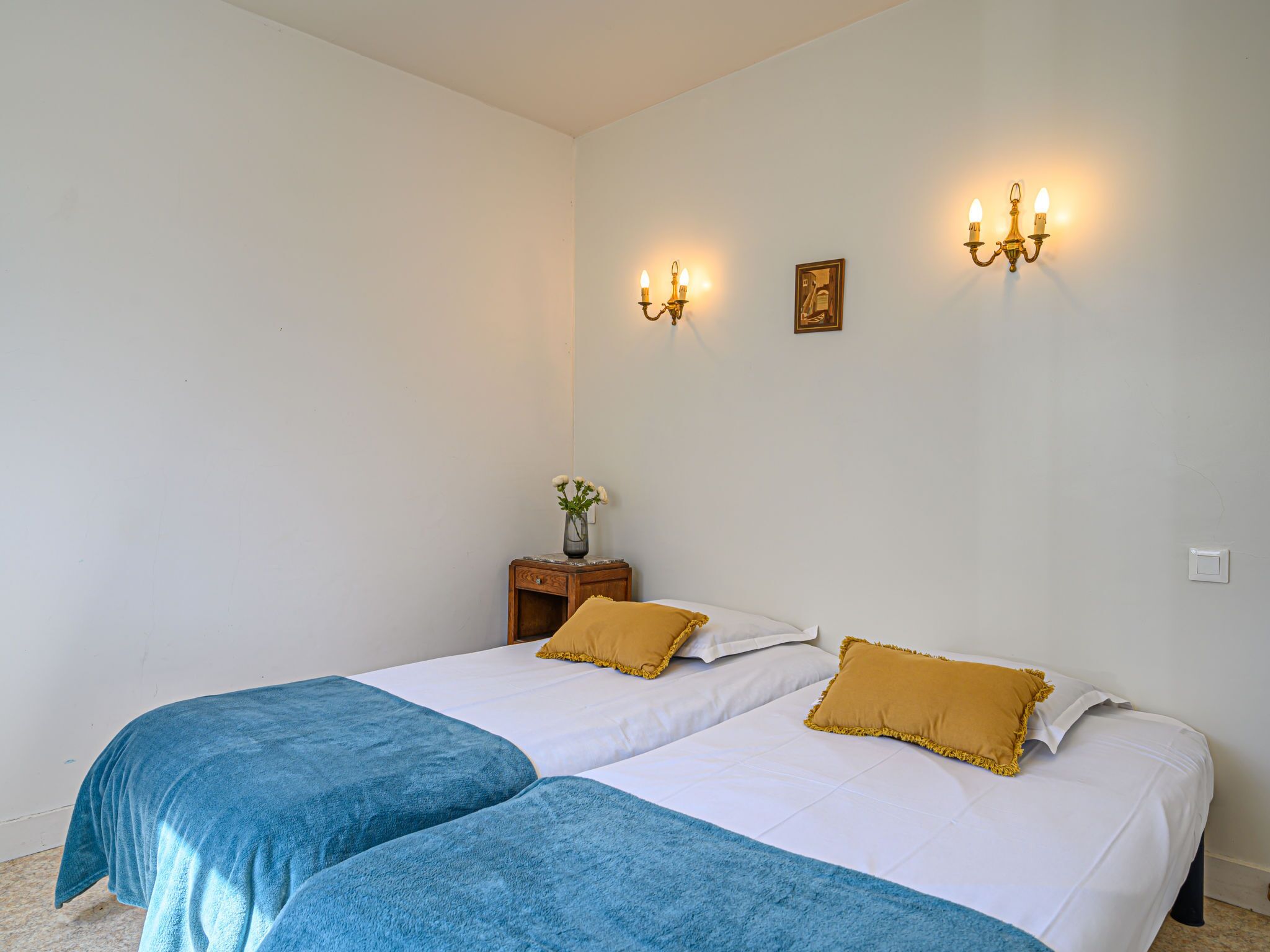 Hotellihuoneessa on kaksi sänkyä, joissa on sinivalkoiset päiväpeitteet, sekä pieni puinen yöpöytä, jonka päällä on maljakko täynnä kukkia.