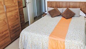 Une chambre d’hôtel avec une armoire en bois, un lit recouvert d’une couvre-lit à motifs floraux et d’un chemin de lit orange, deux oreillers marron et une table de chevet.