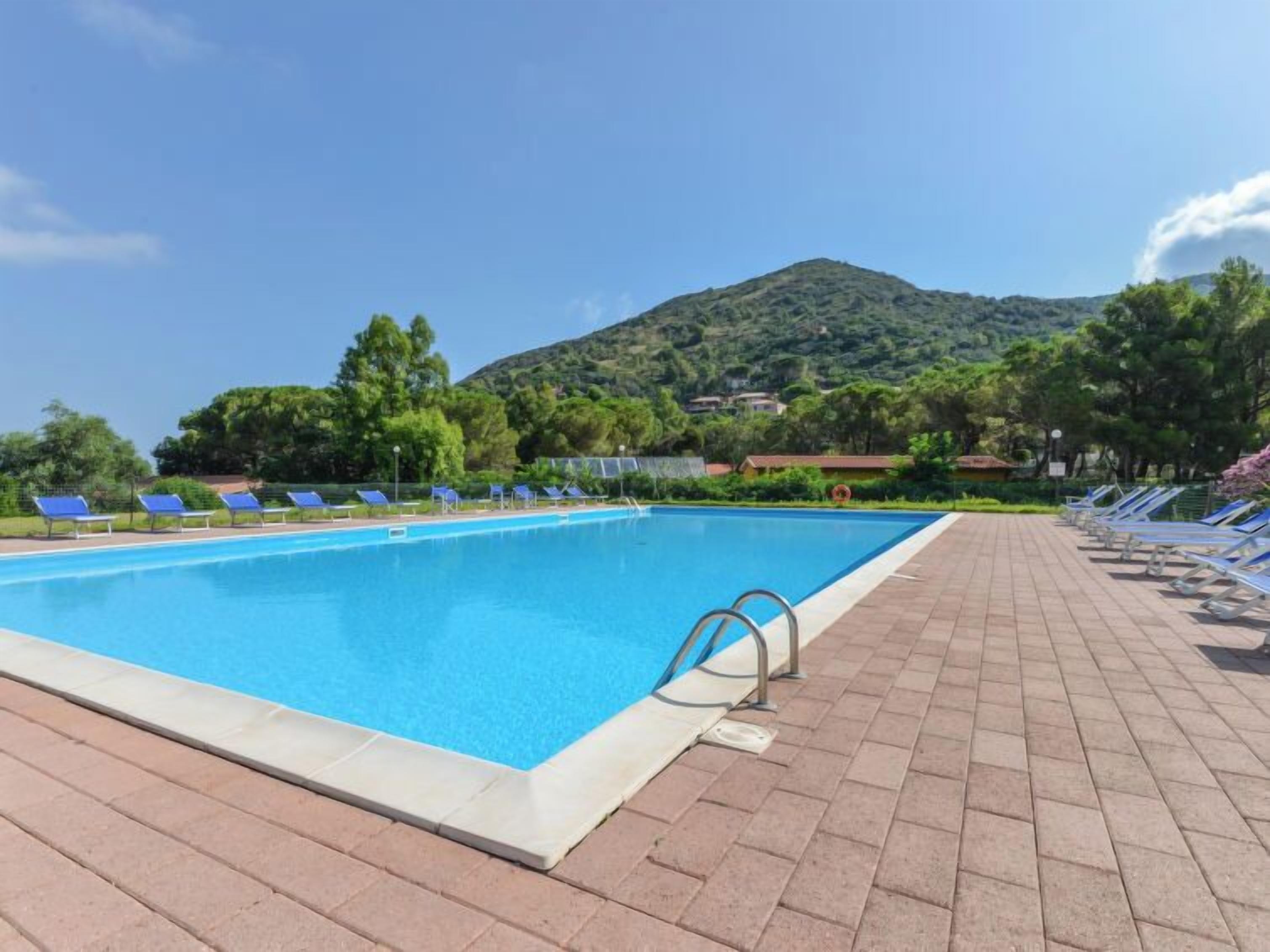 A swimming pool with sun loungers and a paved deck, surrounded by greenery and a hill in the background.