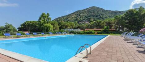 A swimming pool with sun loungers and a paved deck, surrounded by greenery and a hill in the background.