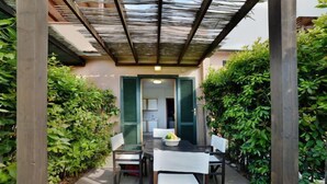 Un patio exterior con una mesa y sillas, rodeado de abundante vegetación y una estructura de pérgola de madera.