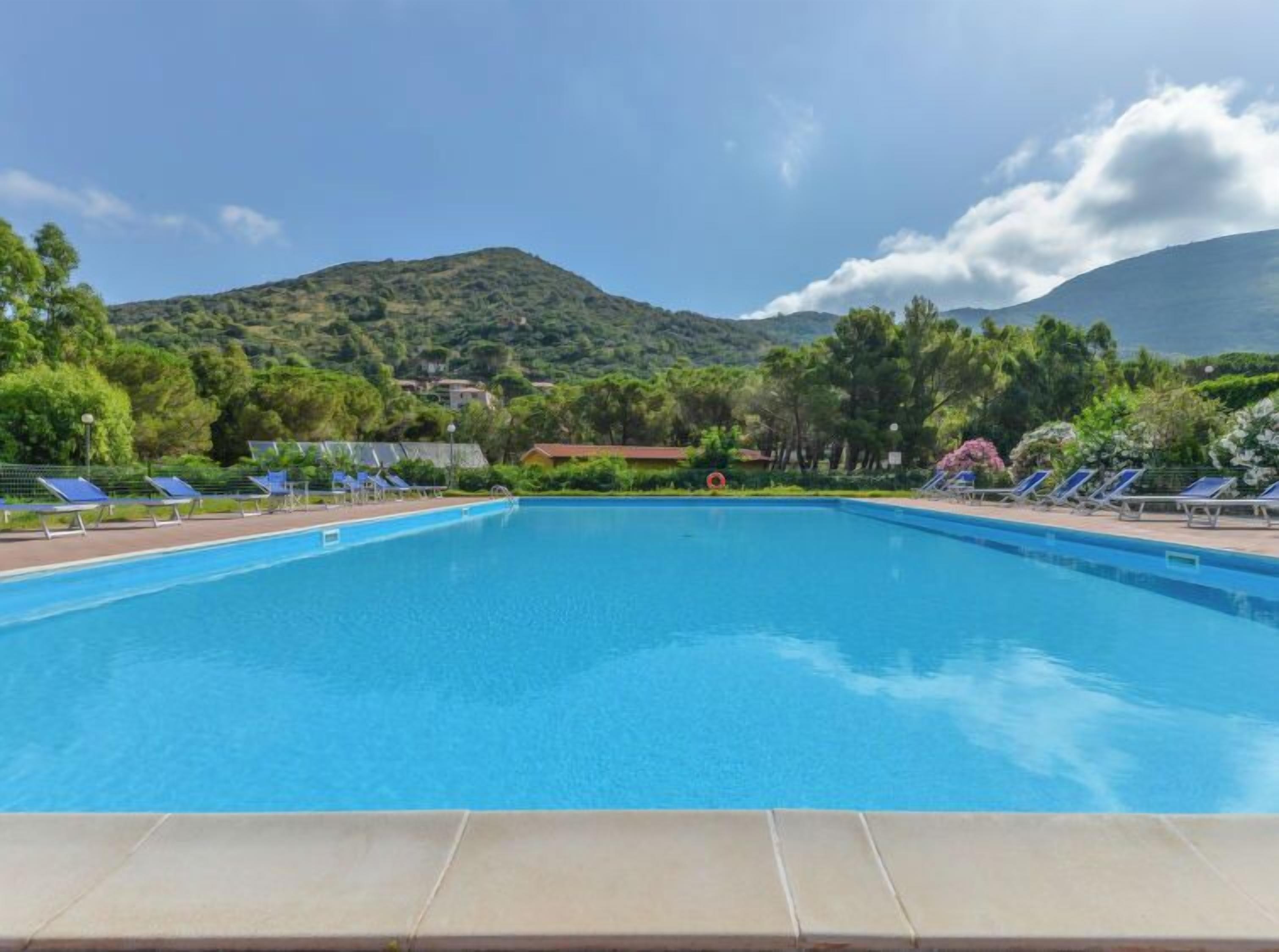 A rectangular swimming pool with clear blue water, surrounded by lounge chairs and a mountainous backdrop.