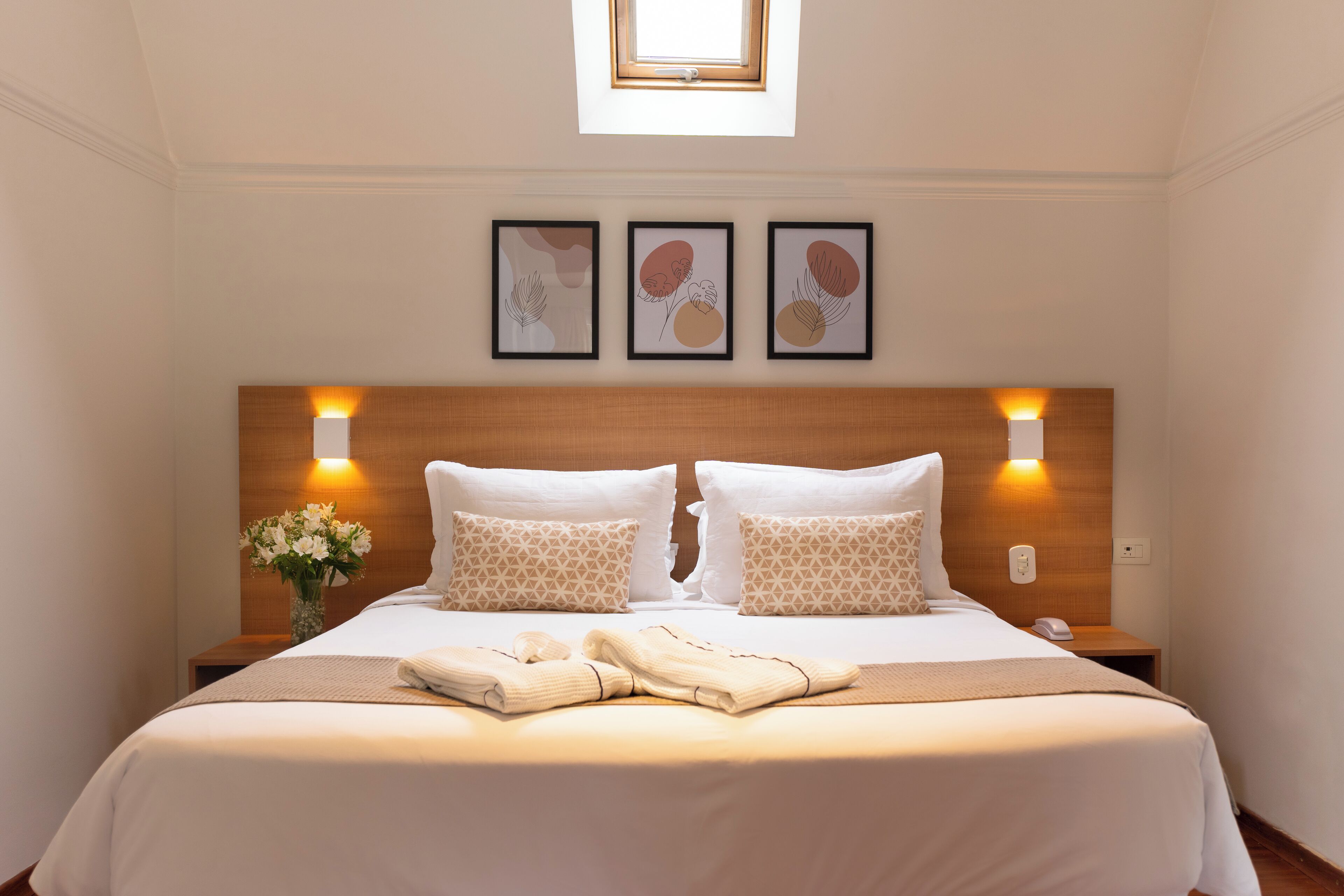 A hotel room with a neatly made bed, a nightstand with a lamp, and three framed artworks on the wall.