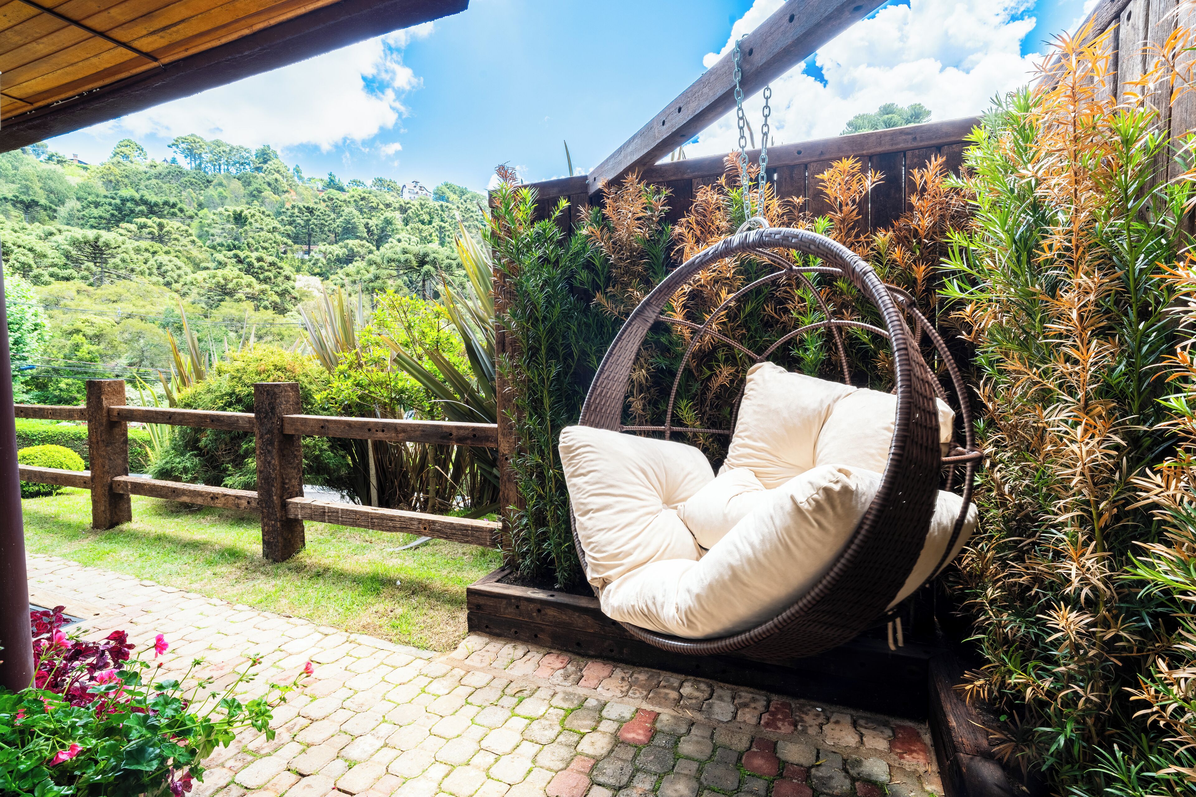 A wicker swing chair with cushions, a wooden fence, and lush greenery in the background.