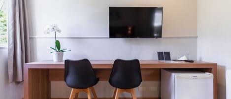 Una habitación moderna con un escritorio de madera, dos sillas negras, un televisor de pantalla plana, un refrigerador pequeño blanco y una orquídea en maceta.