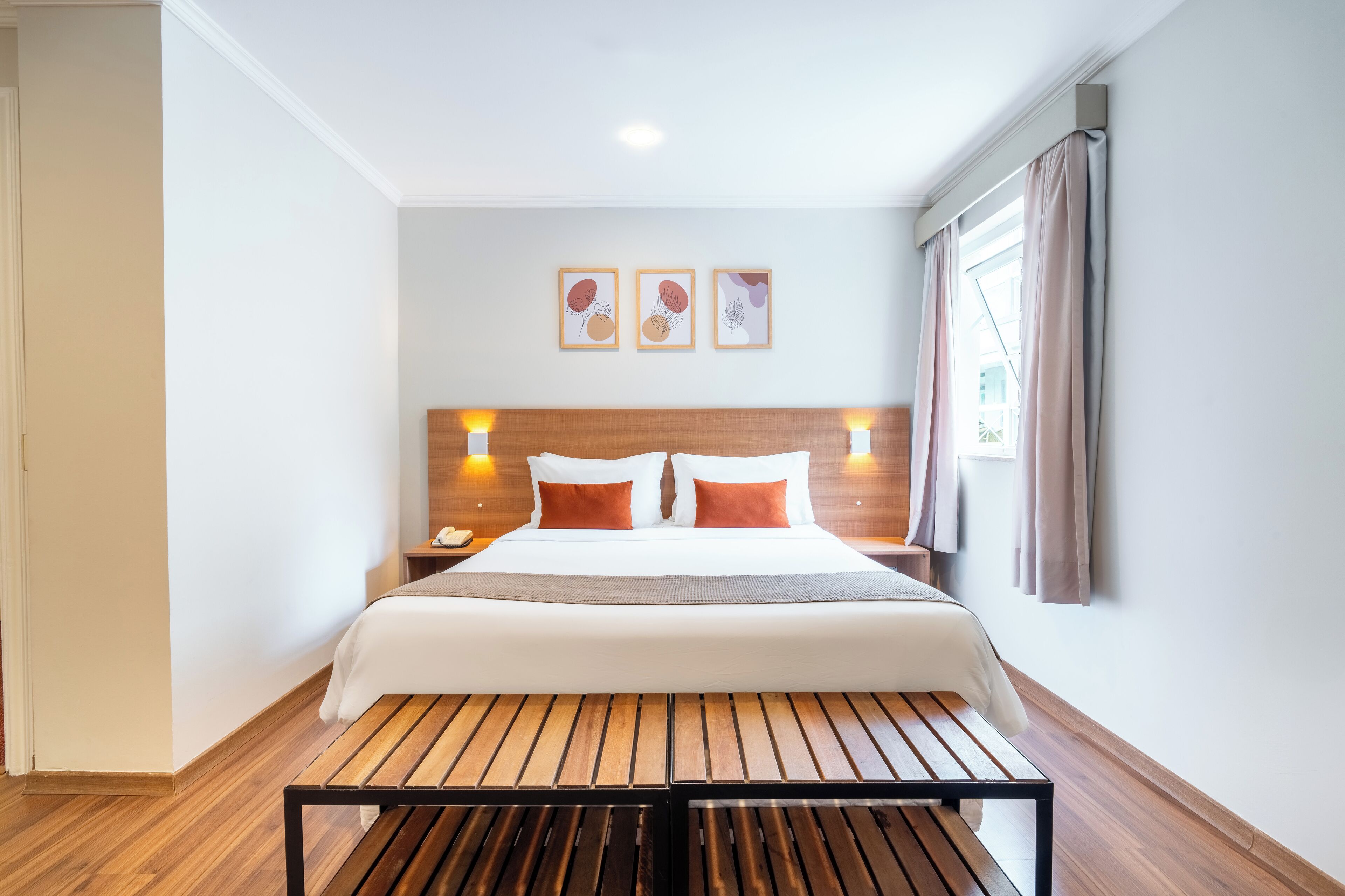 A hotel room with a wooden floor, a bed with white bedding and orange pillows, a nightstand with a phone, and a window with curtains.