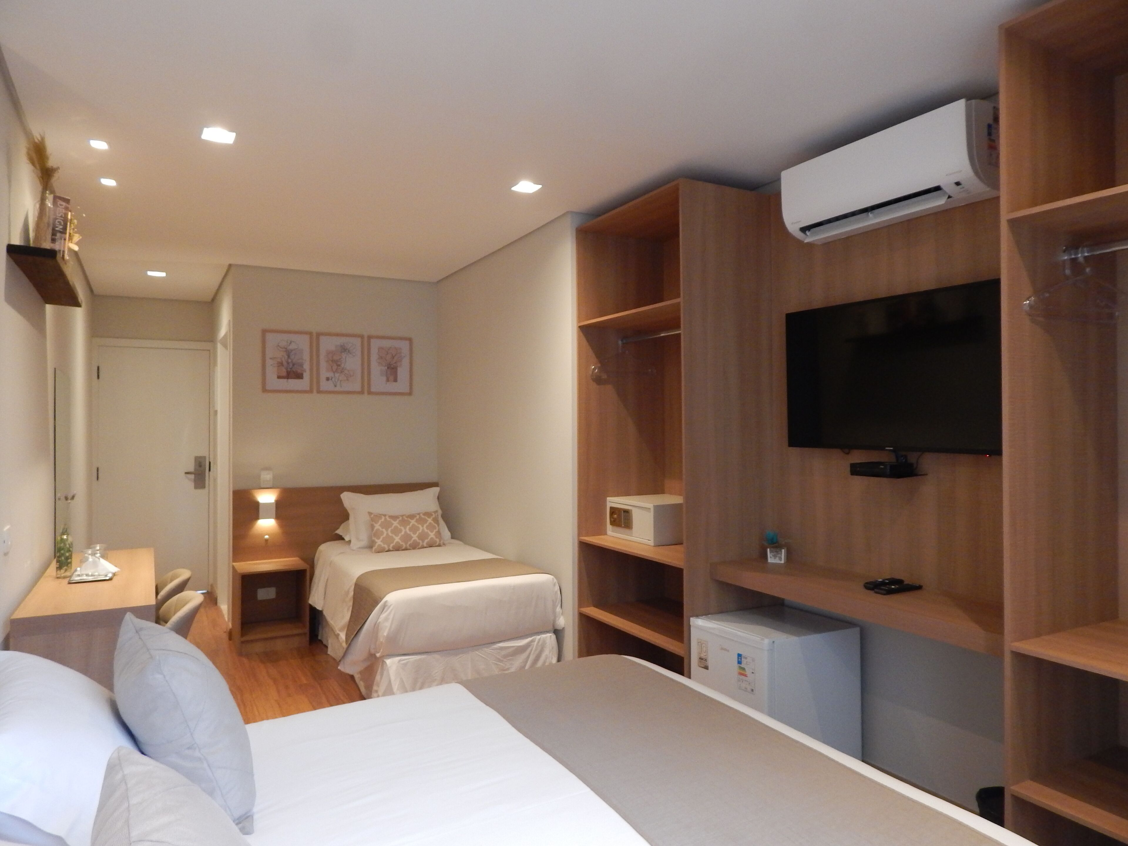Habitación de hotel con una cama grande, un escritorio con silla, un refrigerador pequeño, un televisor montado en la pared y un armario con estantes.