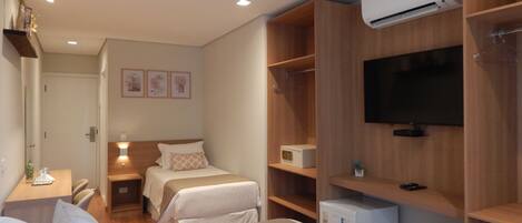 Habitación de hotel con una cama grande, un escritorio con silla, un refrigerador pequeño, un televisor montado en la pared y un armario con estantes.