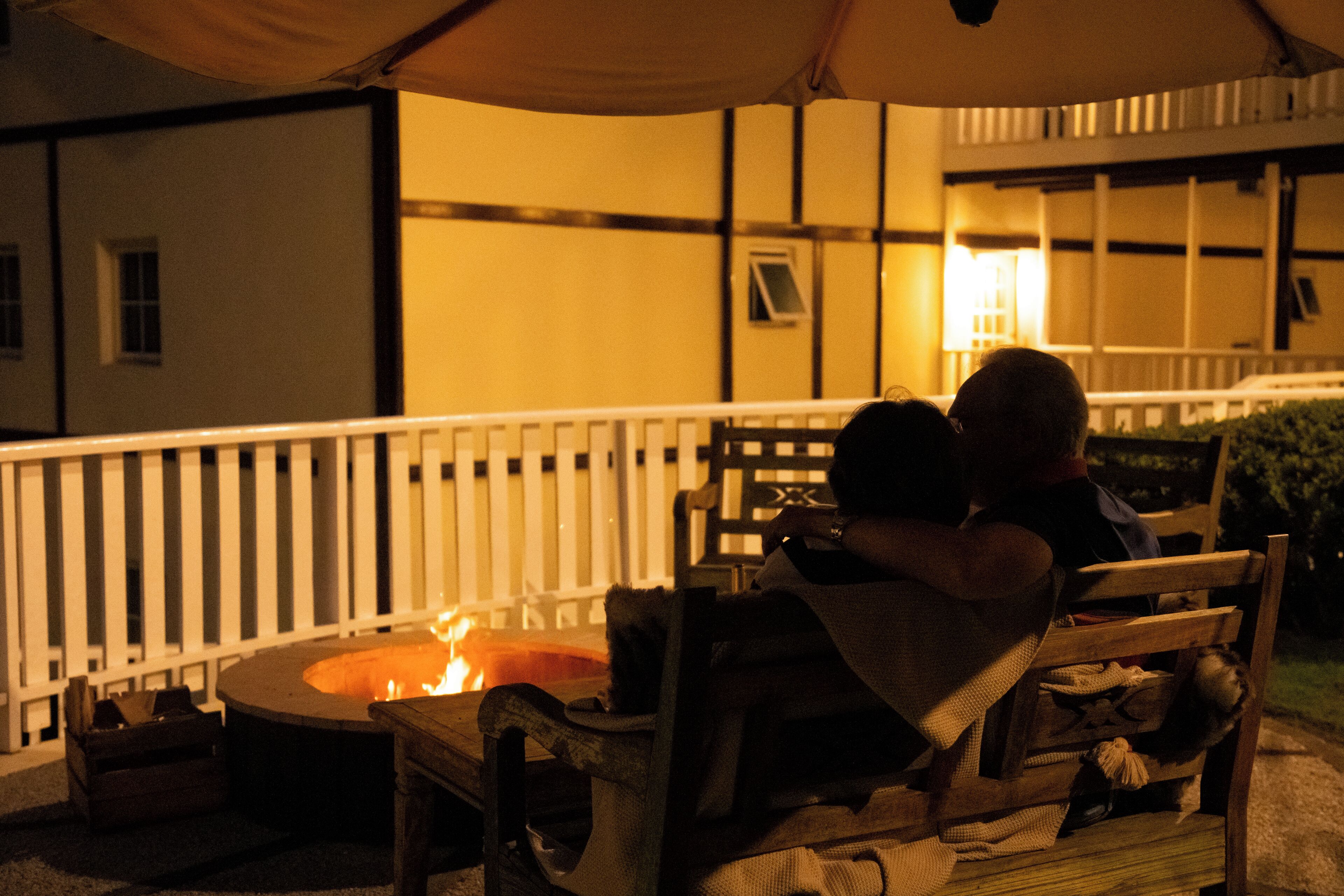 An outdoor seating area with a fire pit, wooden furniture, and a white fence.