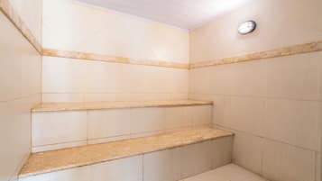 Canto de um quarto com paredes revestidas de azulejos bege e teto branco. Há três degraus bege com um topo semelhante ao mármore. Uma pequena ventilação circular é visível na parede.