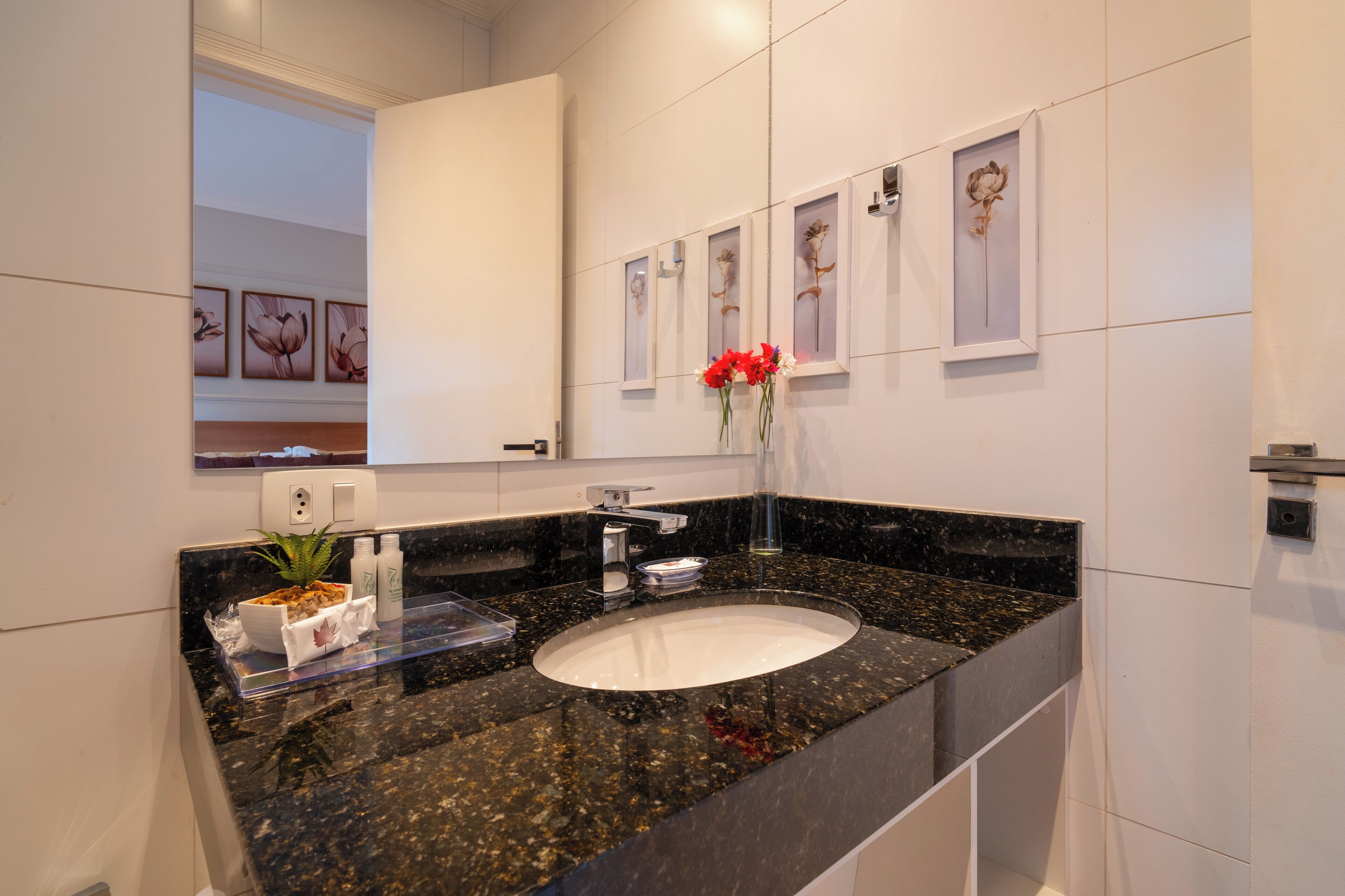 Un baño moderno con encimera de granito, lavamanos blanco y espejo encima. Hay cuadros enmarcados en la pared y un jarrón con flores rojas sobre la encimera.