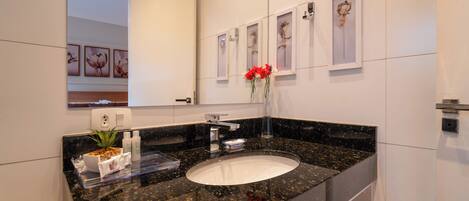 Un baño moderno con encimera de granito, lavamanos blanco y espejo encima. Hay cuadros enmarcados en la pared y un jarrón con flores rojas sobre la encimera.