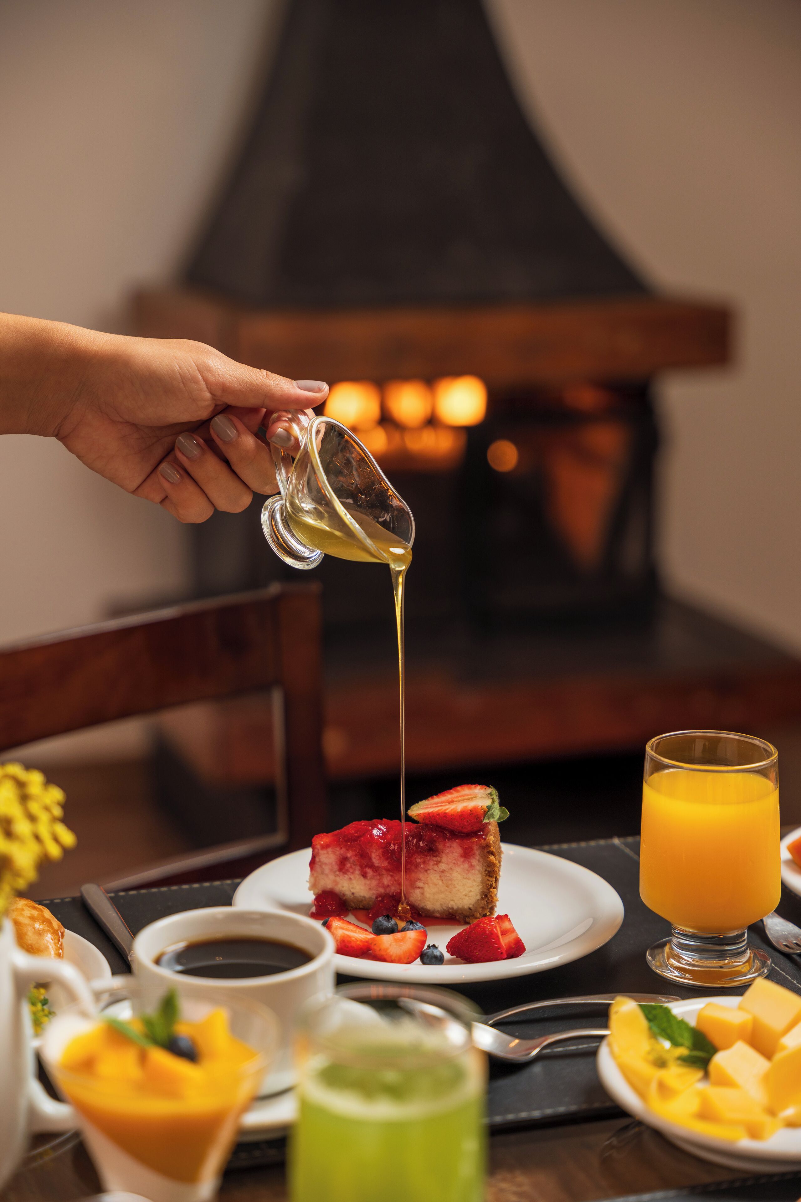 Una escena de desayuno con una mano sirviendo jarabe sobre un postre, un vaso de zumo de naranja y diversas opciones de desayuno sobre una mesa.