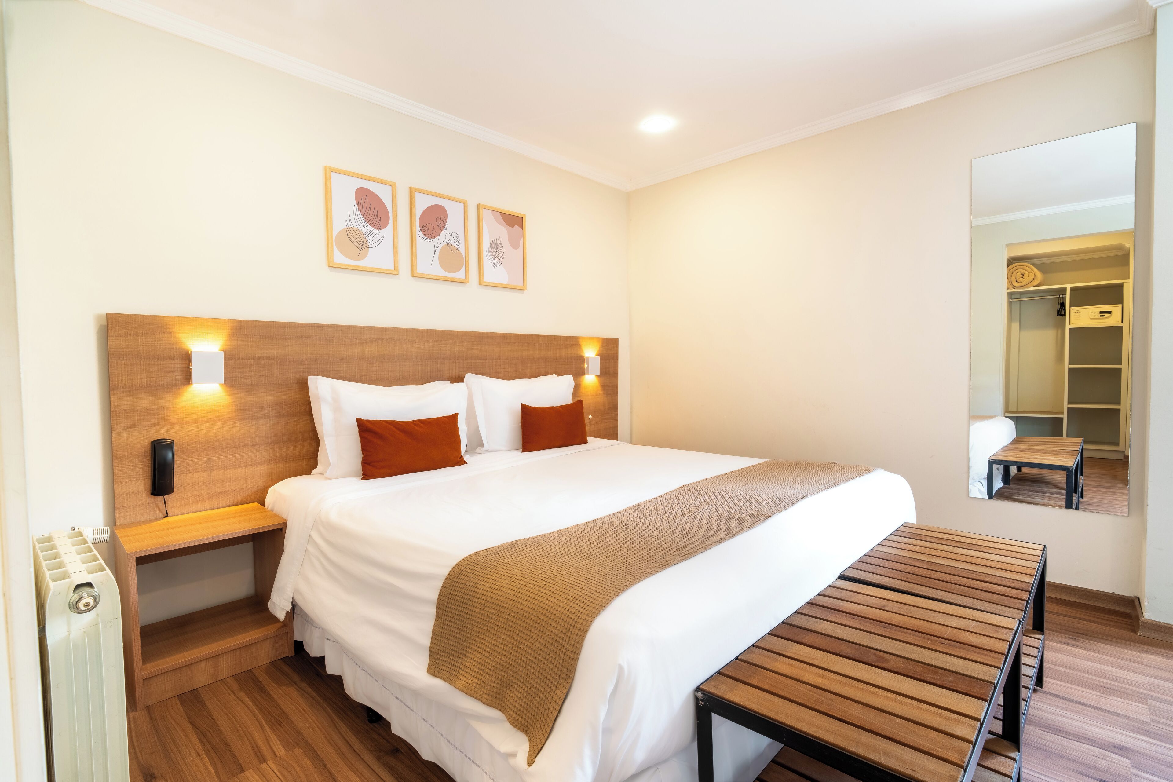 A hotel room with a large bed, wooden headboard, bedside tables, a bench, and a mirror.
