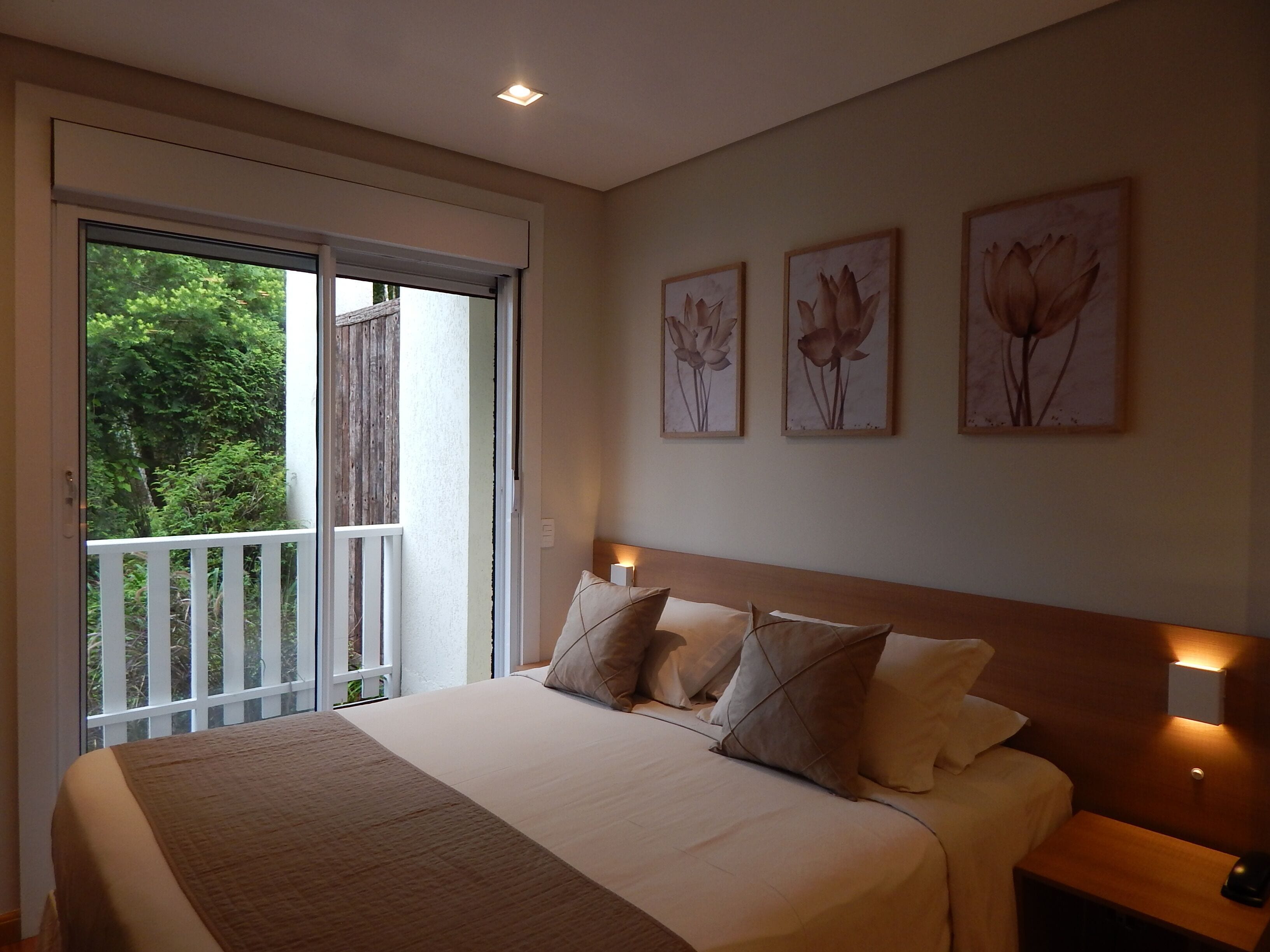 Quarto com cama grande, mesa de cabeceira e luminária na parede. Há três quadros florais emoldurados na parede.
