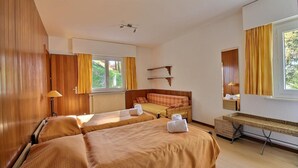 A hotel room with two beds, a window with curtains, a bedside table, a lamp, and a small shelf.