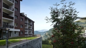 A multi-story building with balconies, a stone wall, and a grassy area with a tree.