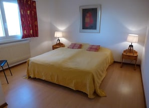 Une chambre avec un grand lit, deux tables de chevet avec des lampes, un radiateur, une fenêtre avec des rideaux et un tableau encadré au mur.
