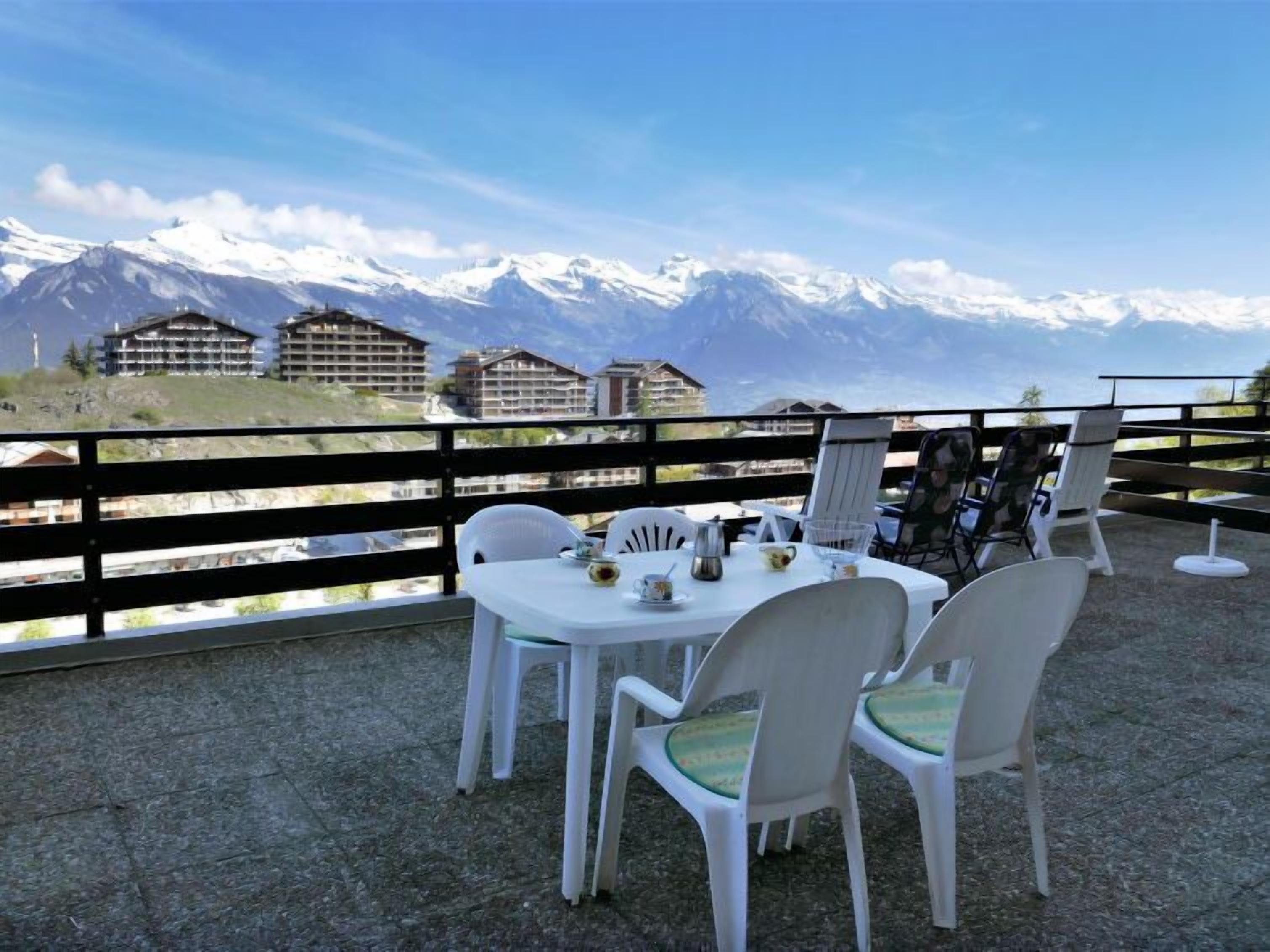 A terrace with white plastic chairs and a table set for dining, overlooking snow-capped mountains and buildings.
