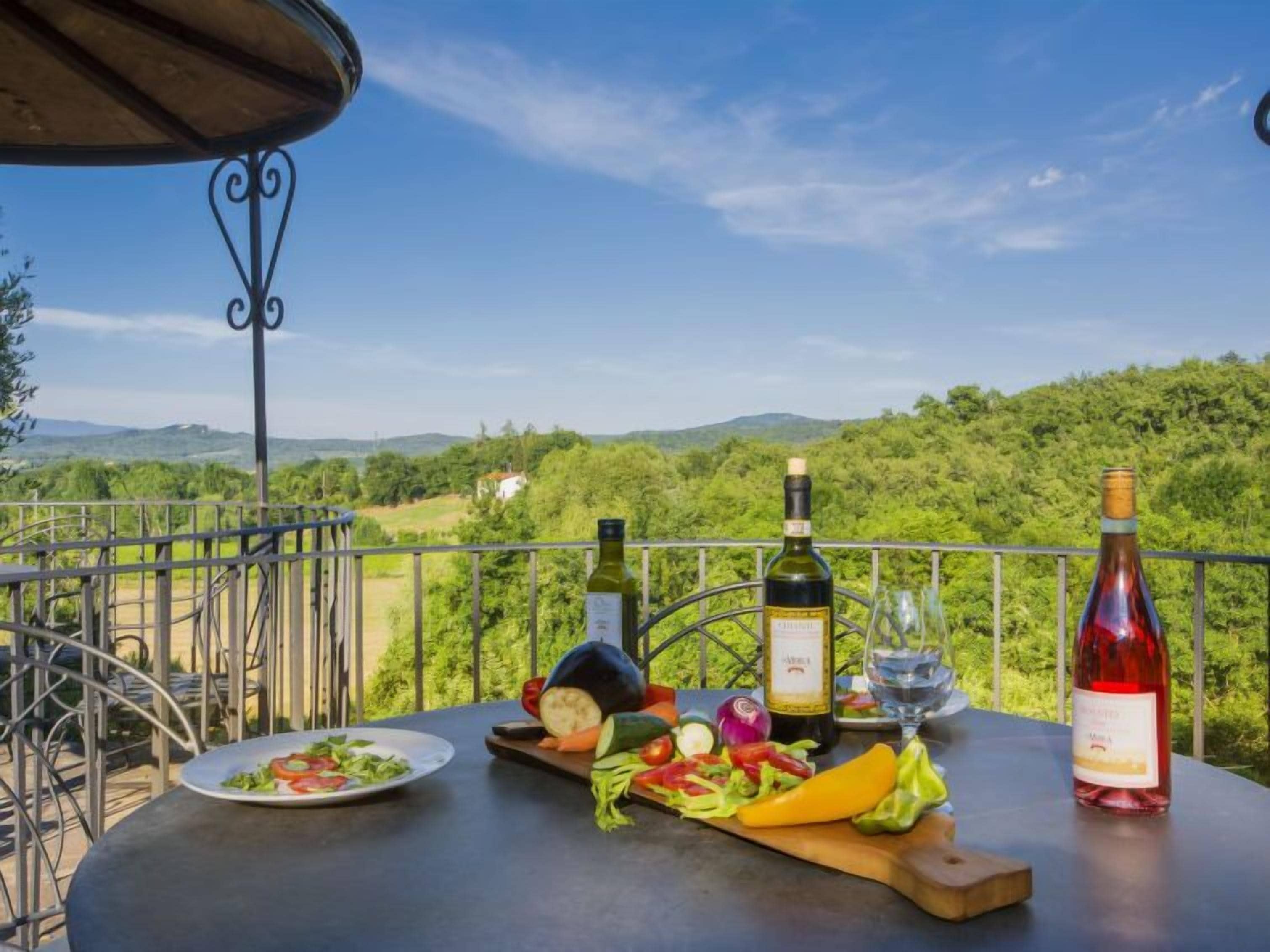 A table with a bottle of wine, a glass, and a plate of salad on a balcony with a scenic view.