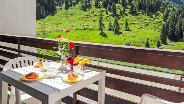 Un balcón con mesa dispuesta para el desayuno, un florero con flores y una vista panorámica de las montañas.