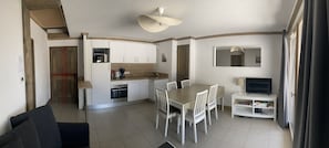 Cocina compacta con armarios blancos, una mesa de comedor con sillas, un televisor sobre un mueble bajo y una lámpara en el techo.