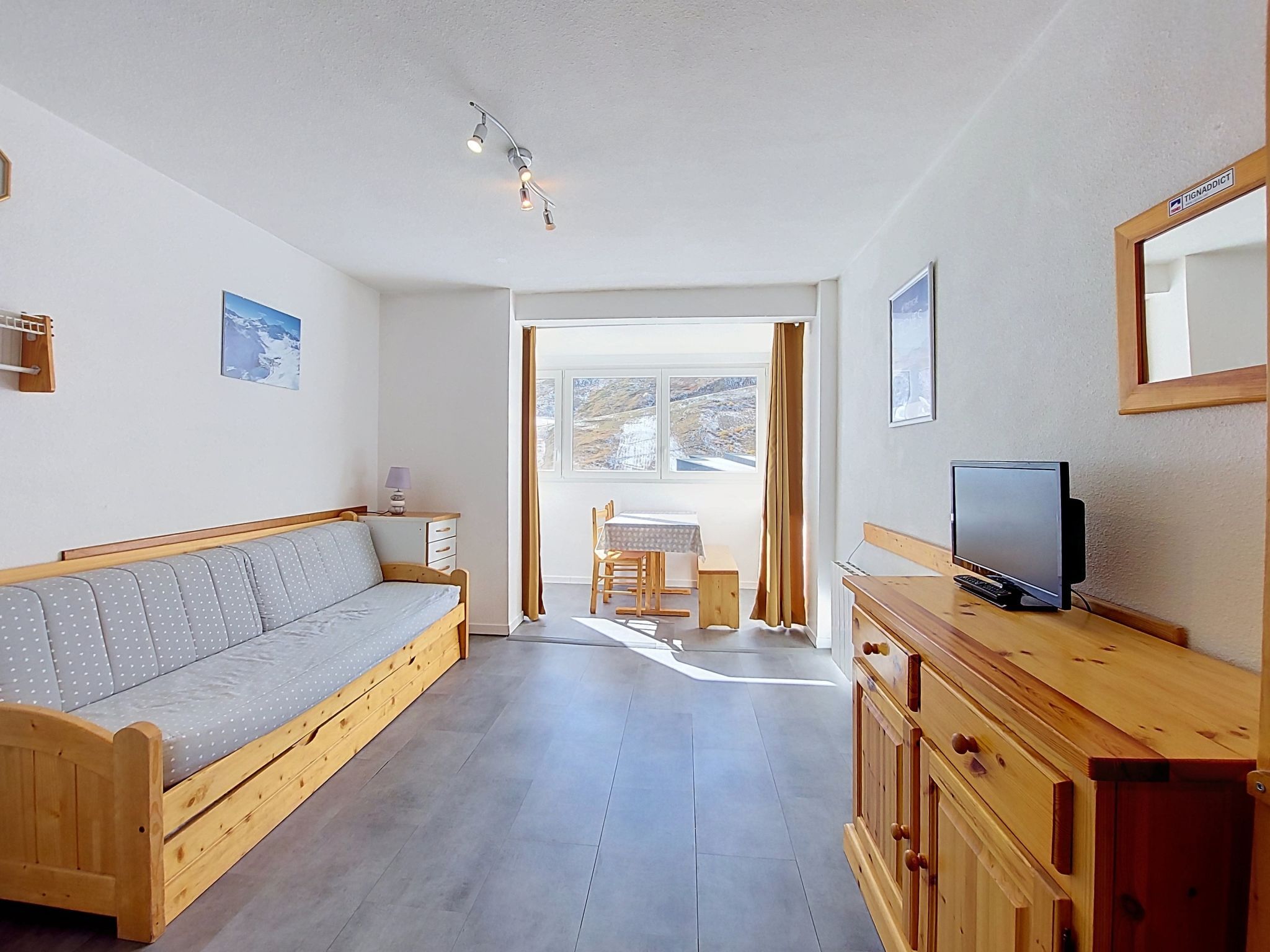 Una habitación con un sofá de madera, un televisor sobre un mueble de madera, una mesa con sillas y una ventana con cortinas.