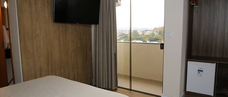A hotel room with a bed, a television, a sliding door to a balcony, and a small refrigerator.